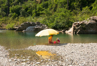 To personer sidder under en gul paraply ved en stenstrand nær en flod i Saint Jean du Gard, Occitanie, Frankrig.