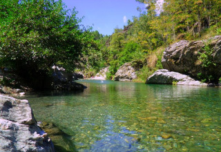 Ein klarer, flacher Fluss, umgeben von Felsen und Bäumen im Bivouac nature Ferienpark, Occitanie, Frankreich.