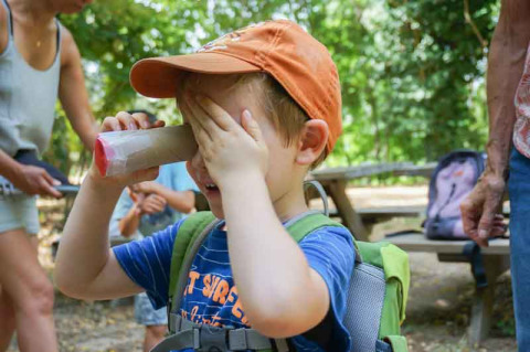 Boy wearing an orange cap plays with a homemade cardboard tube viewer at a nature holiday park in France.