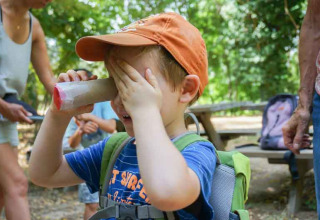 Junge mit orangefarbener Mütze spielt mit selbstgebautem Fernglas im Ferienpark in Occitanie, Frankreich.