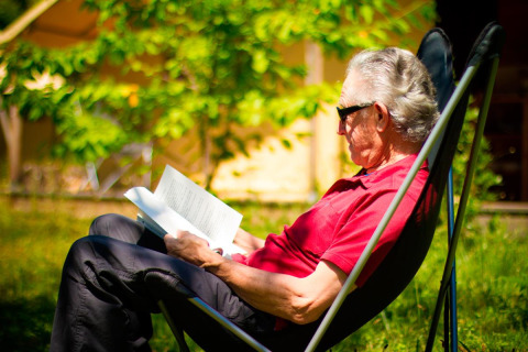 Anziano seduto su una sedia legge un libro all'aperto a Bivouac nature, Occitanie, Francia.