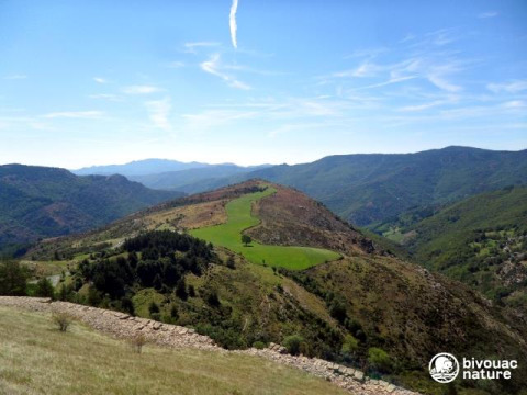 Bjergrigt landskab ved Bivouac Nature feriepark i Occitanie, Frankrig, med grønne marker og blå himmel.