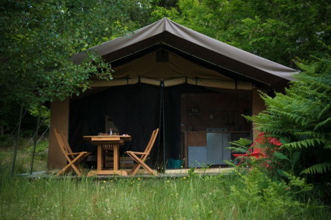 Tenda safari al Bivouac nature in Francia circondata dal verde, con tavolo e sedie in legno davanti.