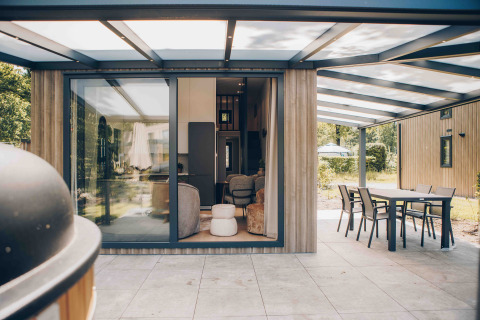Overdekt terras aan een moderne lodge met grote ramen, eethoek buiten en zicht op de tuin.