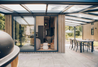 Overdekt terras aan een moderne lodge met grote ramen, eethoek buiten en zicht op de tuin.