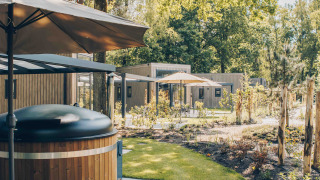 Buitenaanzicht van een lodge met hottub en parasol, omringd door groen gazon en hoge bomen.