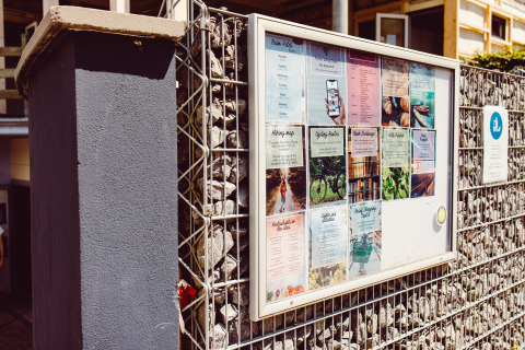 Panneau d'affichage au Camping Liefrange à Wiltz, Luxembourg, avec flyers et cartes sous verre sur un mur en pierre.