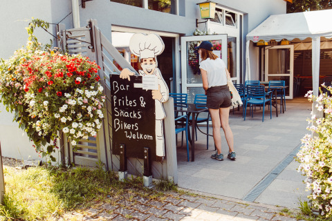 Woman at the entrance of a campsite café with flowers, tables and a sign, Camping Liefrange, Luxembourg.