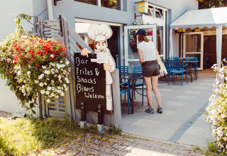 Mujer en la entrada de un café en el camping con flores, mesas y cartel, Camping Liefrange, Luxemburgo.