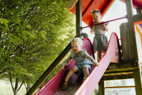 Two small children playing on a slide under green trees at Camping Liefrange holiday park in Wiltz, Luxembourg.