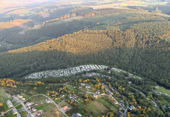Vista aerea di un glamping circondato da colline boscose, case e piazzole per tende e roulotte sottostanti.