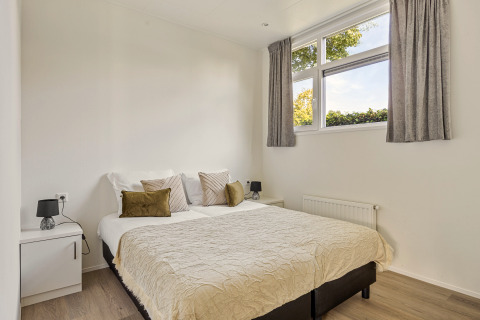 Chambre lumineuse avec lit double, murs blancs et grandes fenêtres dans une lodge à Recreatiepark het Esmeer.