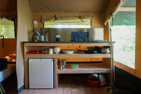Coin cuisine dans la tente Baroudeur avec réchaud à gaz, mini-frigo, ustensiles et étagères en bois.