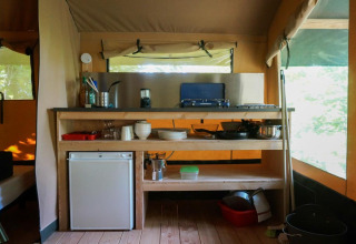 Küchenbereich im Baroudeur-Zelt mit Gasherd, Minikühlschrank und Küchenutensilien auf Holzregalen.