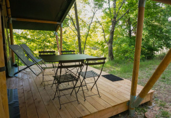 Terrazza in legno della tenda safari Baroudeur con tavolo, sedie e sdraio immersa nel verde del bosco.