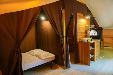 The «Canadienne» safari tent at Bivouac nature in France, showing a bed, wooden furniture, and curtains.