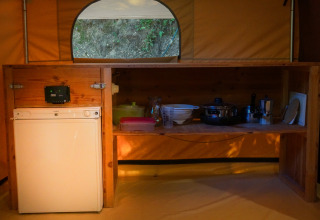 Kampeerkeuken met houten rek, kleine koelkast en keukengerei in een safaritent in Frankrijk.