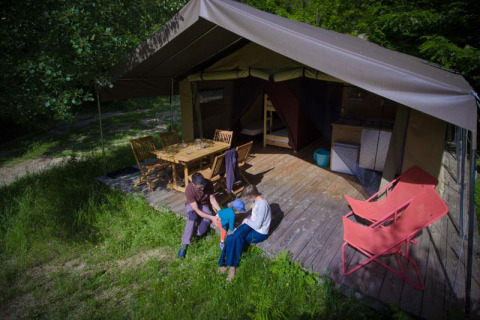 Famiglia si rilassa davanti a una tenda safari con terrazza in legno, tavolo, sedie e prato verde.