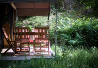Træmøbler på verandaen af en safaritelt ved Bivouac nature, omgivet af frodig grøn vegetation i Frankrig.