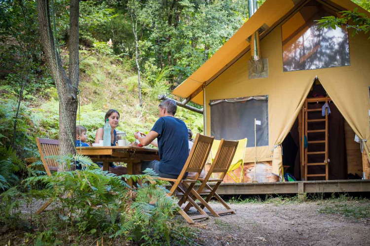 Familia comiendo al aire libre frente a una tienda safari Cabane en Bivouac nature, Francia, rodeados de bosque.