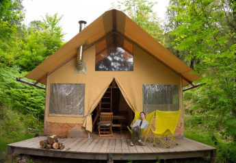 Vrouw zit ontspannen op gele stoelen voor safaritent Cabane, omgeven door weelderig bos.