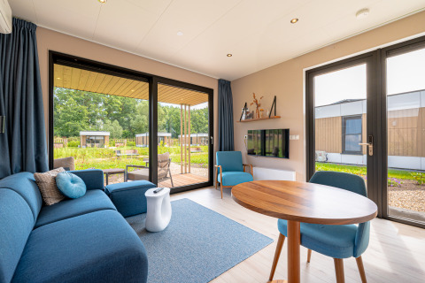 Lyst og moderne opholdsområde i Water lodge 2 med blå sofa, spisebord og udsigt til naturen udenfor.