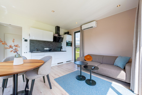 Intérieur de lodge moderne avec cuisine ouverte, canapé gris, tapis bleu, tables rondes et espace repas.