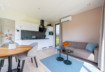 Moderne lodge met open keuken, grijze bank, blauw vloerkleed, ronde tafels en eethoek met tuinzicht.