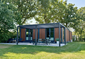 Et moderne Forest Cube-lodge med træfacade og udendørs terrasse, omgivet af grønne træer og græs.