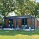 Et moderne Forest Cube-lodge med træfacade og udendørs terrasse, omgivet af grønne træer og græs.