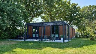 Lodge Forest Cube moderne à façade bois, grande terrasse extérieure et pelouse verdoyante entourée d’arbres.