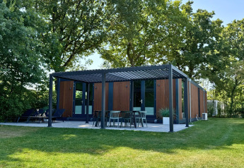 Lodge Forest Cube moderno con fachada de madera, terraza al aire libre y césped rodeado de árboles altos.