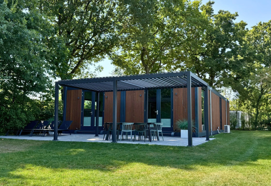 Un lodge Forest Cube moderno con esterno in legno, terrazza ombreggiata e giardino verde tra gli alberi.