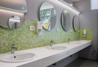 Salle de bains commune moderne avec miroirs ronds, carrelage vert et plusieurs lavabos dans un parc en Croatie.