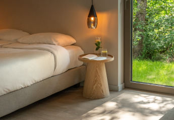 Dormitorio minimalista de cabaña con cama doble, mesa de madera, lámpara colgante y ventana al bosque.