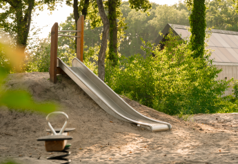 Aire de jeux avec toboggan en métal et jouet sur ressort près d’un lodge dans une zone boisée et ensoleillée.