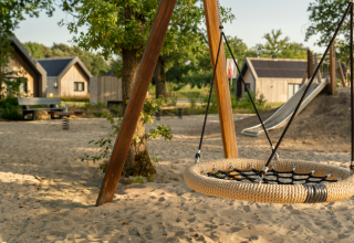 Kinderspielplatz mit Nestschaukel und Rutsche auf Sand neben Lodges in grüner Umgebung.