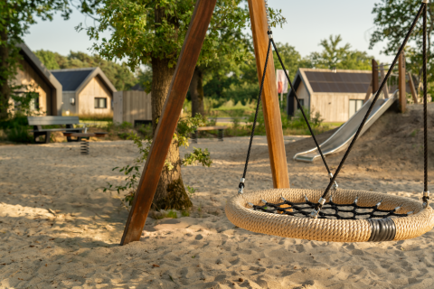 Børnelegeplads med gynge og rutsjebane på sandgrund ved hytter i grønne omgivelser på en lodge.