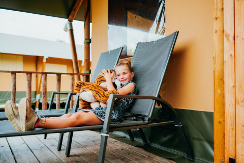 Mädchen entspannt sich auf Liegestuhl mit Tigerstofftier vor Luxus-Safarizelt im Holiday Park Sallandshoeve.