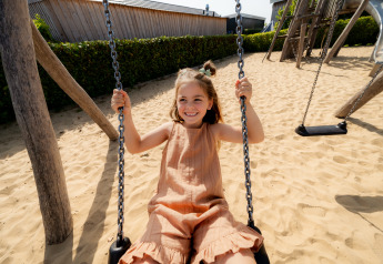 Girl swings on a sandy playground at a lodge, enjoying a sunny day in a joyful outdoor setting.