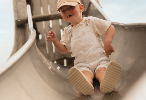 Un niño pequeño con peto claro y gorra desciende por un tobogán de metal en Biebosch, Resort Mooi Bemelen, Países Bajos.