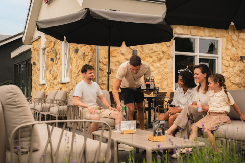 Fem personer, herunder et barn, nyder en hyggelig eftermiddag på en terrasse ved en lodge med stenmur.