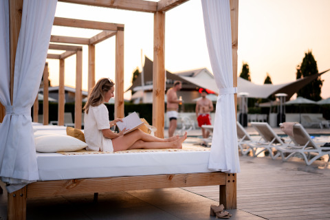 Woman reading a book on a canopy daybed by the pool at Biebosch Lodge, Resort Mooi Bemelen, Netherlands.
