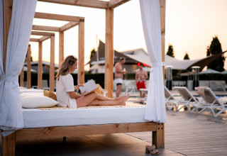 Woman reading a book on a canopy daybed by the pool at Biebosch Lodge, Resort Mooi Bemelen, Netherlands.