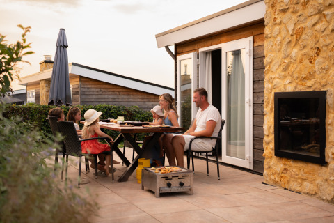 Een familie eet samen buiten op het terras van een gezellige lodge onder de avondzon.