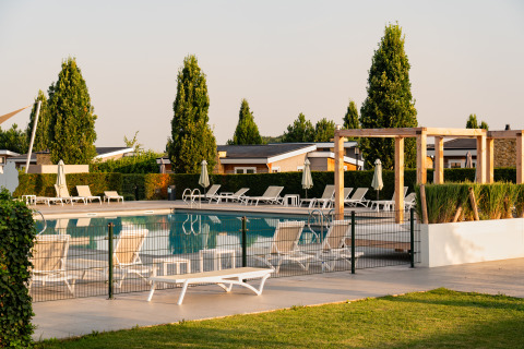 Udendørs swimmingpool ved en lodge med liggestole, parasoller og grønne træer en solrig dag.