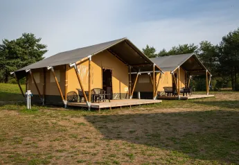 Deux tentes safari modernes avec terrasses et chaises, placées sur un terrain herbeux entouré d’arbres.