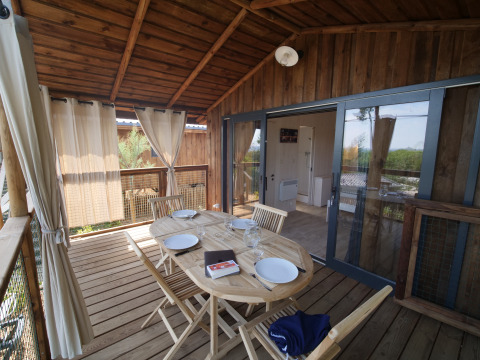 Porche de madera en una cabaña, mesa preparada para cuatro personas con sillas, cortinas y vistas.