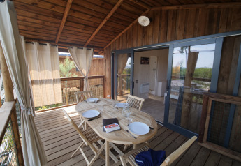 Veranda in legno di un lodge con tavolo apparecchiato per quattro, sedie e tende, vista sulla natura.