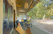 Überdachte Terrasse eines Safarizelts mit Sitzmöbeln und Blick auf einen waldreichen Campingplatz.